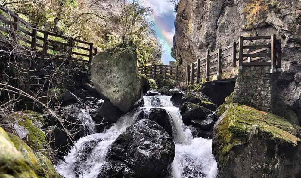 Un torrente scorre tra rocce e passerelle con parapetti in legno.