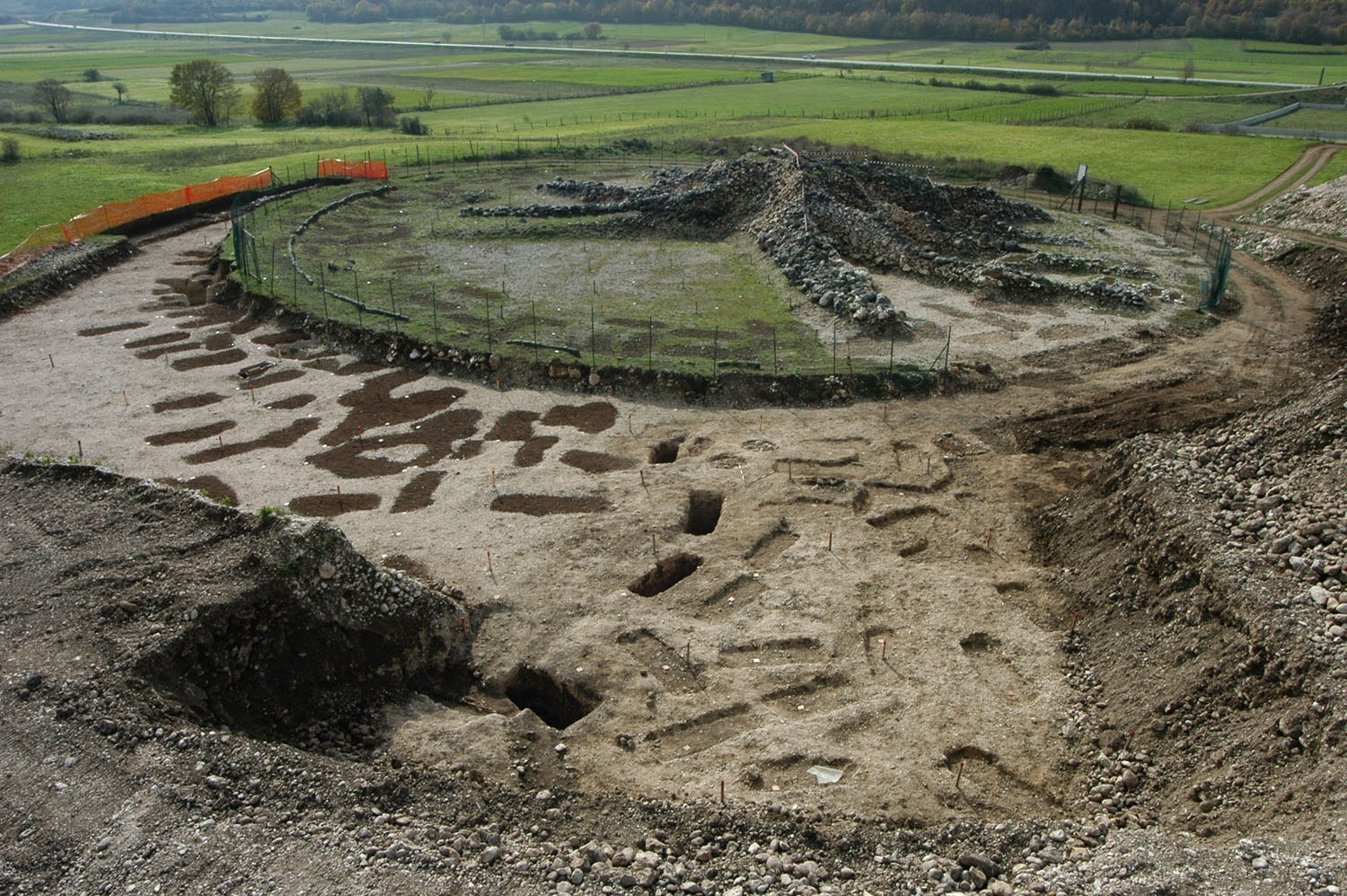 Un’area di scavo archeologico con strutture circolari in pietra e fosse nel terreno.